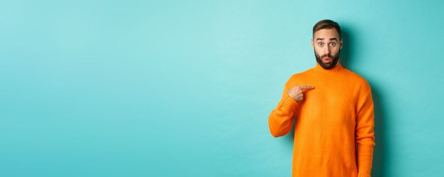 Man pointing at himself with surprise face, being chosen, standing confused against light blue background.