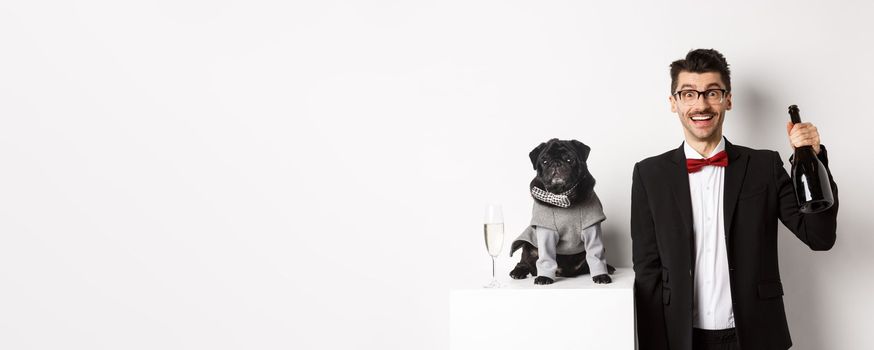 Pets, winter holidays and New Year concept. Cheerful man with cute black pug dog celebrating Christmas party, holding champagne bottle and smiling, white background.