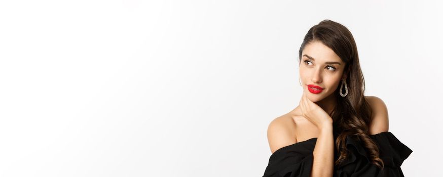 Fashion and beauty concept. Elegant woman with earrings and black dress, looking left sensual, standing over white background. Copy space