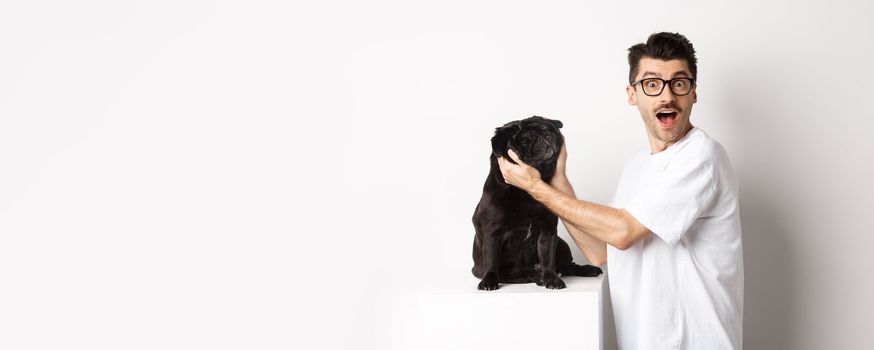 Happy young man showing you cute face of his pug. Dog owner loving his pet, standing over white background.