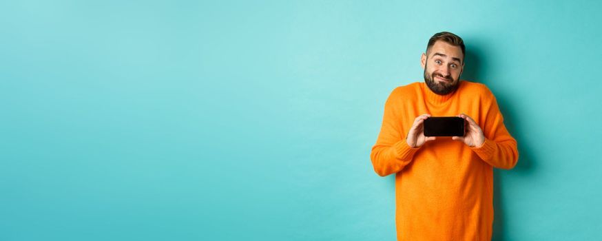 Clueless guy shrugging and showing mobile screen, indecisive emotion, standing in orange sweater over turquoise background.