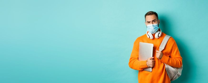 Handsome caucasian man with headphones and backpack, holding laptop and wearing medical mask, looking doubtful and skeptical, standing over light blue background.