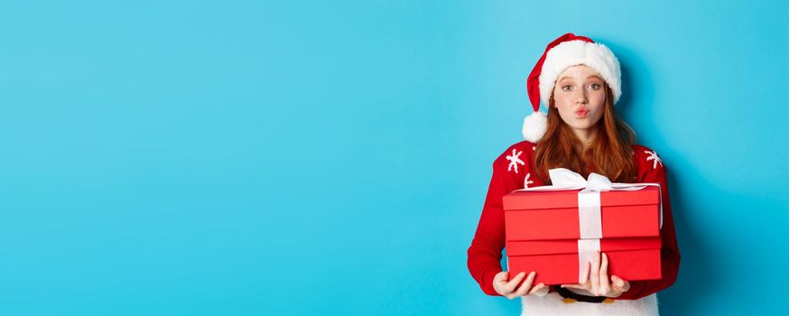 Happy holidays and Christmas concept. Cute redhead girl holding presents and pucker lips for kiss, wearing santa hat and funny sweater, blue background.