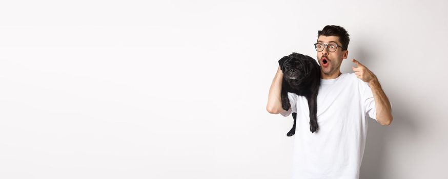 Amazed young man holding cute black dog on shoulder, pointing finger left at promo offer, staring impressed and speechless, standing over white background.