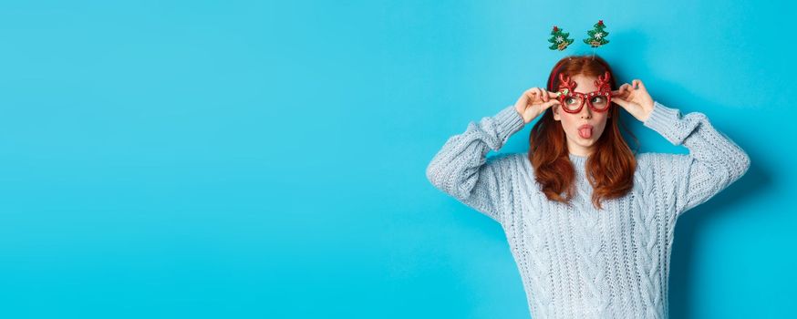 Christmas party and celebration concept. Silly redhead girl enjoying New Year, wearing funny glasses and headband, showing tongue and staring left at logo, blue background.