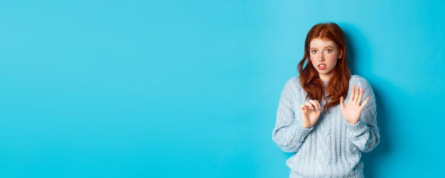 Worried redhead girl refusing or declining an offer, shaking hands and looking anxiously at camera, rejecting something unpleasant, standing over blue background.