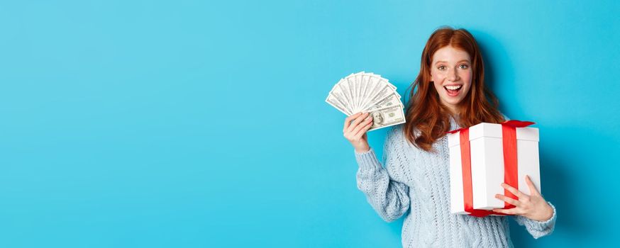 Christmas and shopping concept. Happy redhead woman holding money and big xmas present, showing dollars and gift, smiling pleased, standing over blue background.