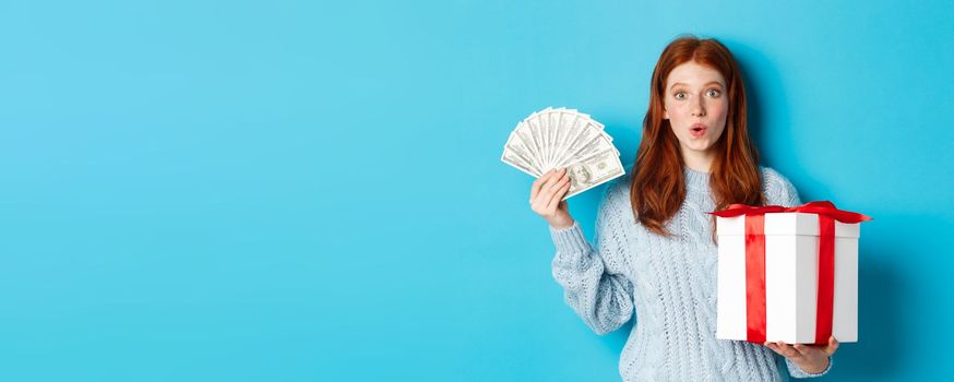 Christmas and shopping concept. Excited redhead girl looking at camera, holding big New Year gift and dollars, buying presents, standing over blue background.