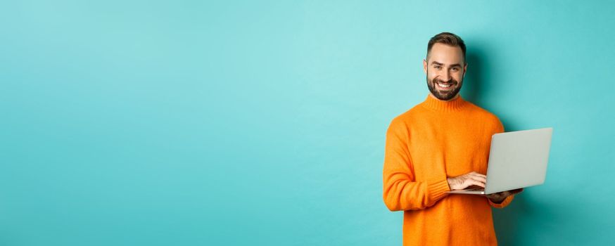 Handsome adult man freelancer working with laptop, smiling at camera, typing on computer keyboard, standing over light blue background.