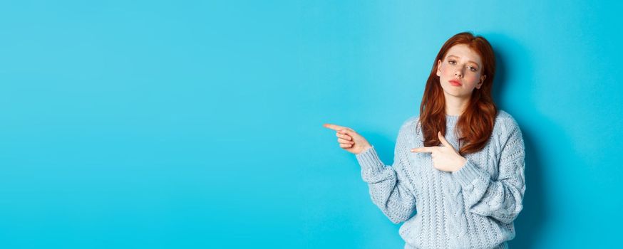 Moody teenage girl with red hair, pointing fingers left at logo, staring bothered and bored, standing over blue background.