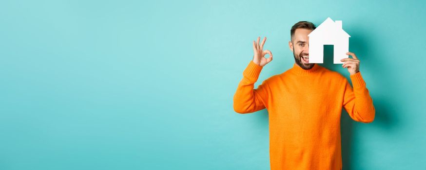 Real estate concept. Satisfied man recommending agency, showing okay sign and house maket, standing over blue background.