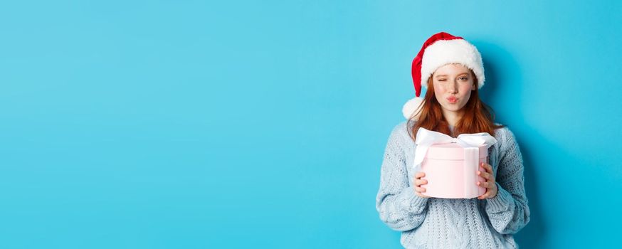 Winter holidays and Christmas Eve concept. Sassy redhead girl in sweater and Santa hat, holding New Year gift and looking at camera, standing against blue background.