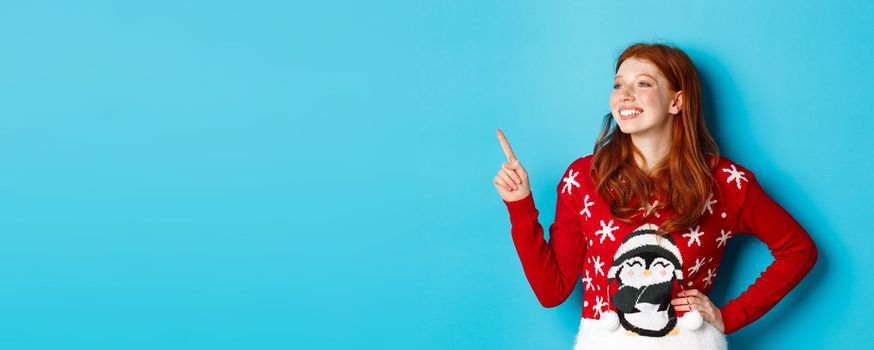 Winter holidays and Christmas Eve concept. Happy and cute redhead girl in xmas sweater, looking and pointing at upper left corner amazed, standing over blue background.