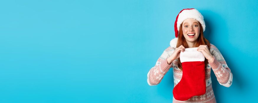 Beautiful redhead girl in santa hat open Christmas stocking and looking surprised, receiving xmas gift, standing over blue background.