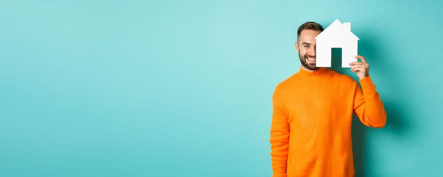 Real estate concept. Happy young man searching for home rent, holding house paper maket and smiling, standing over blue background.