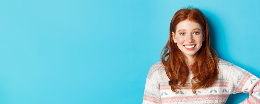 Close-up of attractive female student with red hair, wearing winter sweater, smiling happy and staring at camera, blue background.