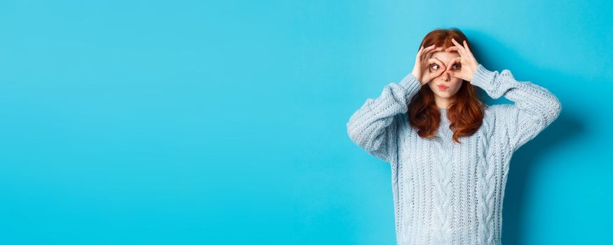 Funny redhead female model in sweater, staring at camera through fingers glasses, express interest and amazement, standing over blue background.