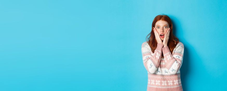 Shocked and surprised redhead girl staring at camera in awe, express complete disbelief, standing against blue background.