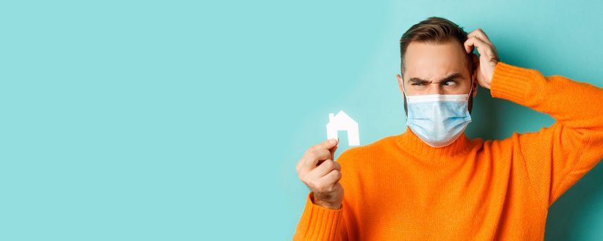 Real estate and coronavirus pandemic concept. Confused man in face mask scartching head while looking at small paper house, wearing orange sweater near light blue studio background.