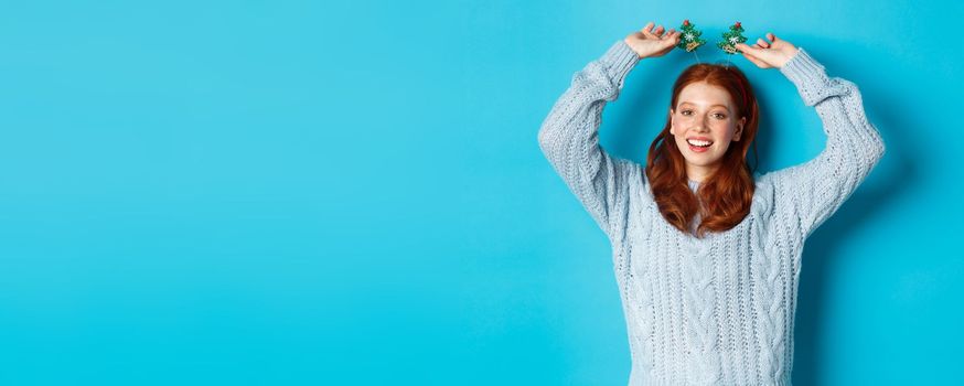 Winter holidays and Christmas sales concept. Beautiful redhead female model celebrating New Year, wearing funny party headband and sweater, smiling at camera.