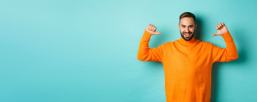 Handsome confident man pointing at himself, looking self-assured, standing in orange sweater against light blue background.
