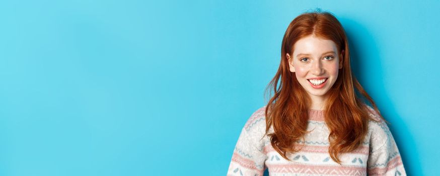 Close-up of cute redhead girl in sweater smiling happy at camera, standing against blue background.