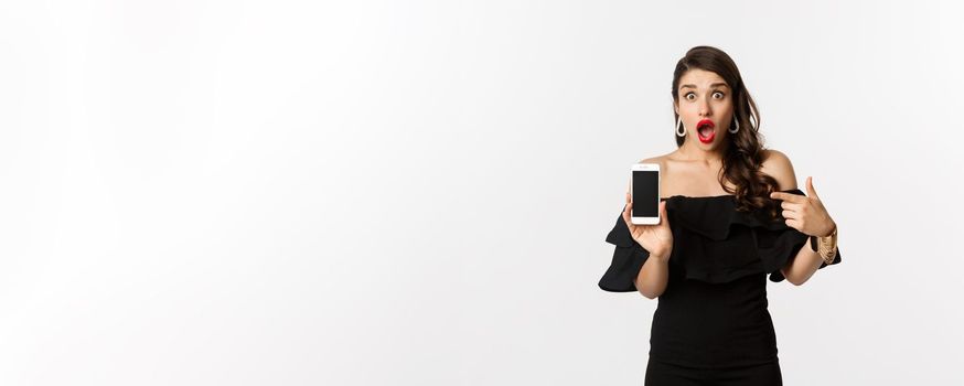 Online shopping concept. Fashionable woman in black dress pointing finger at smartphone screen, showing application, standing over white background.