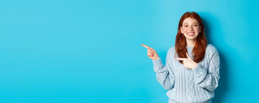 Cute teenager girl with red hair and freckles, pointing fingers left at logo and smiling, showing advertisement, standing over blue background.