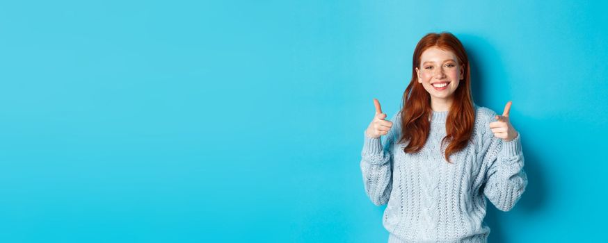 Cheerful teen girl with red hair, pointing fingers at camera and smiling, congratulating or praising you, standing over blue background.