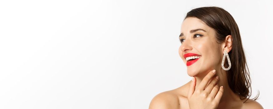 Fashion and beauty concept. Headshot of gorgeous brunette woman with red lipstick, earrings, laughing and looking left, standing over white background.