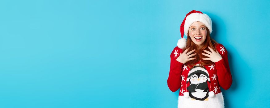 Happy holidays and Christmas concept. Surprised redhead girl receiving unexpected gift, gasping amazed and staring with plesant disbelief, standing in santa hat against blue background.