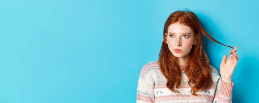 Close-up of thoughtful pretty redhead girl looking left, playing with hair strand and pondering, standing in winter sweater against blue background.