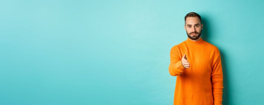 Confident man extending hand for handshake, greeting you, looking self-assured, standing over light blue background. Copy space