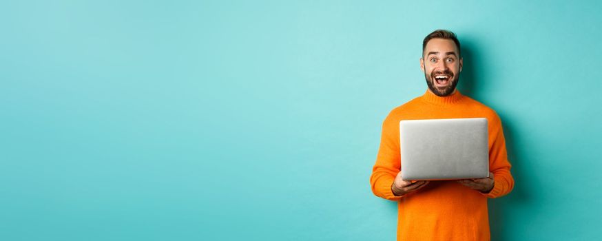 Happy man using laptop and looking excited at camera, standing with computer against light blue background.