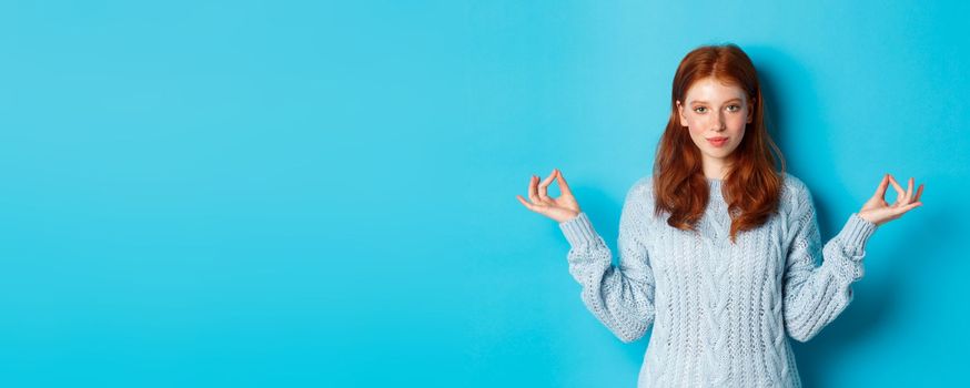 Smiling confident girl with red hair staying patient, holding hands in zen, meditation pose and staring at camera, practice yoga, standing calm against blue background.