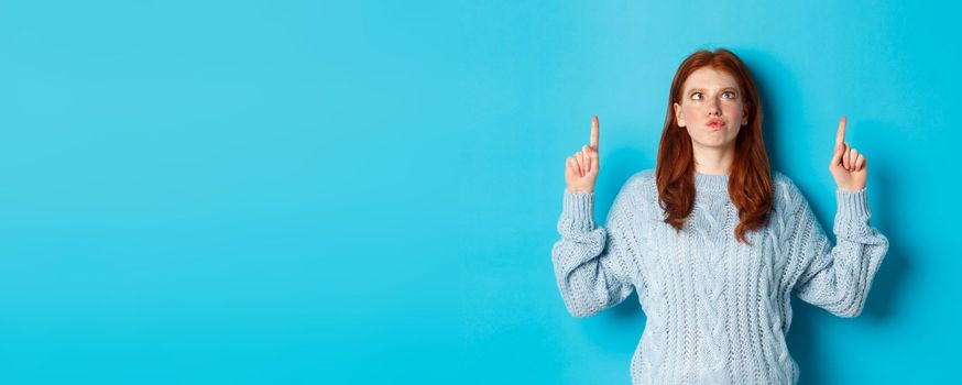 Winter holidays and people concept. Funny and silly redhead girl in sweater, pointing fingers up and squint eyes, standing over blue background.