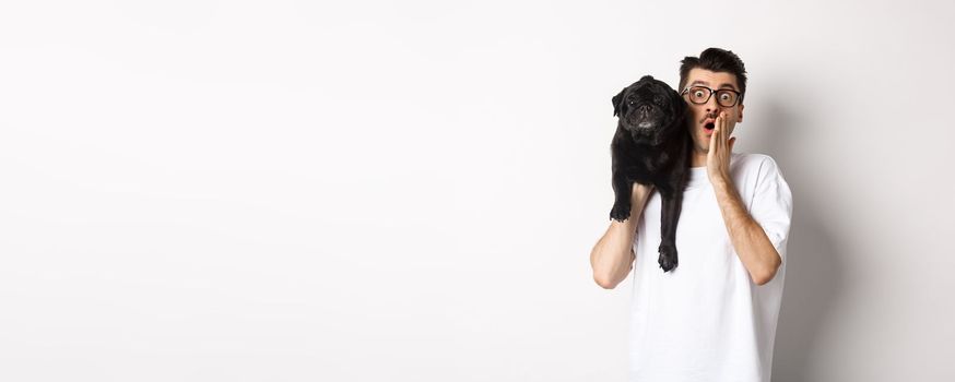 Image of shocked dog owner staring at camera and gasping impressed, cute black pug sitting on his shoulder and staring at you, white background.