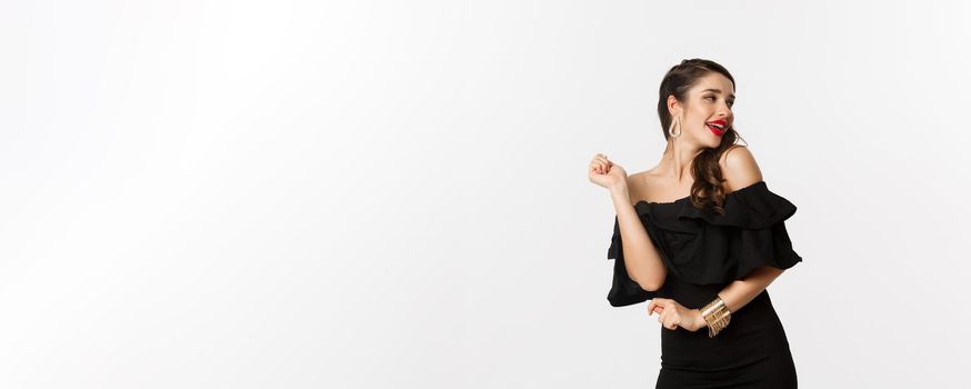 Fashion and beauty. Woman celebrating and dancing in black dress, having fun and smiling, standing over white background.