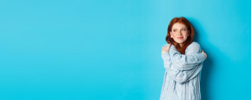 Happy cute redhead girl hugging herself, wearing comfortable and warm sweater, smiling at camera, standing over blue background.