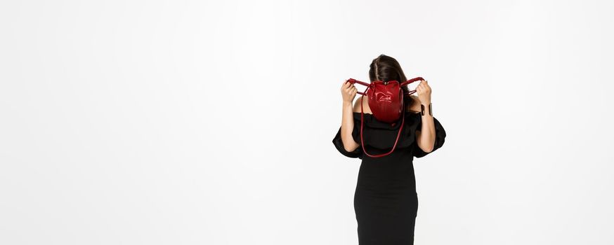 Beauty and fashion concept. Full length of young woman sticking head inside purse and searching something, wearing black dress and high heels, standing over white background.