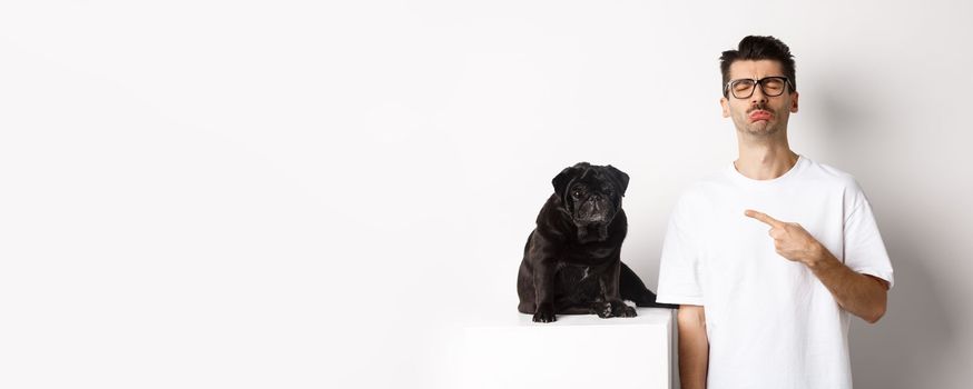 Sad and gloomy pet owner pointing at his black pug dog and sobbing, standing upster against white background.