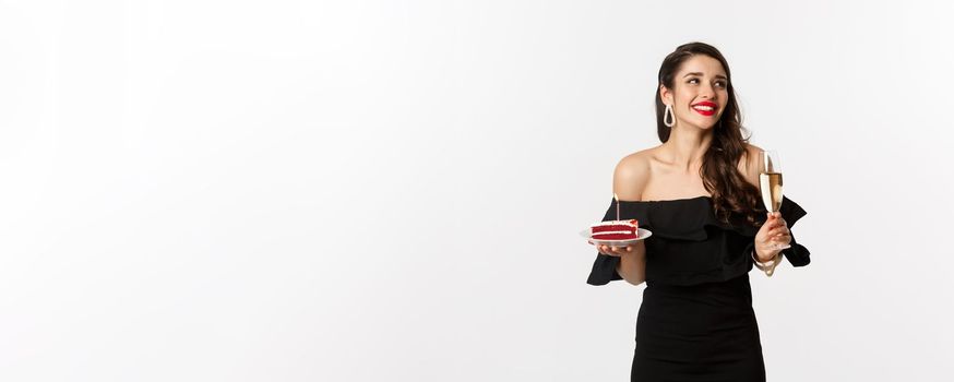 Celebration and party concept. Fashionable woman holding birthday cake with candle and drinking champagne, smiling and looking aside, standing over white background.