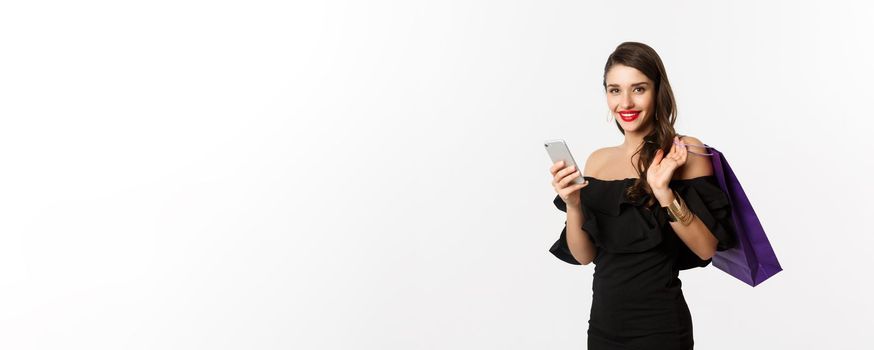 Beauty and shopping concept. Gorgeous woman in elegant black dress and makeup, order online on mobile phone, holding bag and smiling, standing over white background.