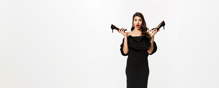 Beauty and fashion concept. Full length of excited glamour woman in black dress, showing high heels and looking excited, dressing up for party, white background.