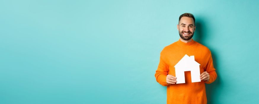 Real estate concept. Happy young man searching for home rent, holding house paper maket and smiling, standing over blue background.