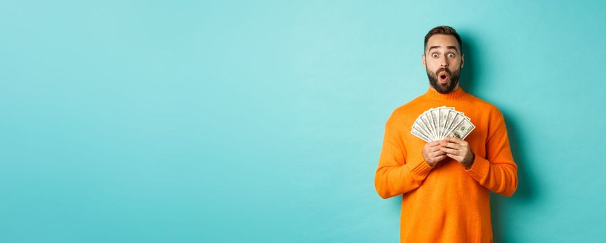 Photo of surprised guy holding money, looking amazed, standing with dollars against turquoise background. Copy space