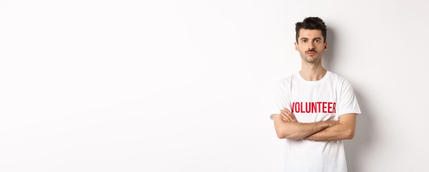 Serious young male volunteer in white t-shirt, holding arms crossed on chest, looking at camera, ready to help.