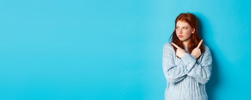 Indecisive redhead teenage girl making decision, pointing fingers sideways and looking left doubtful, standing in sweater against blue background.