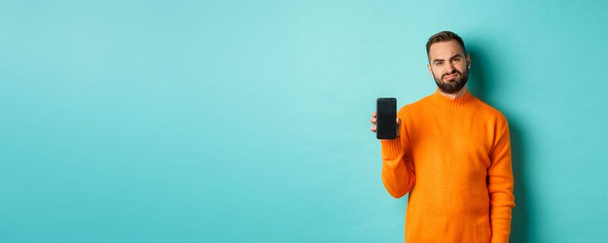 Displeased and disappointed man in orange sweater, grimacing and showing mobile screen, standing over light blue background.