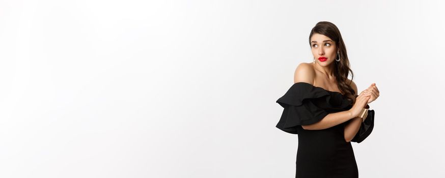 Fashion and beauty. Young silly woman in black dress pouting and looking timid, standing in black glamour dress over white background.